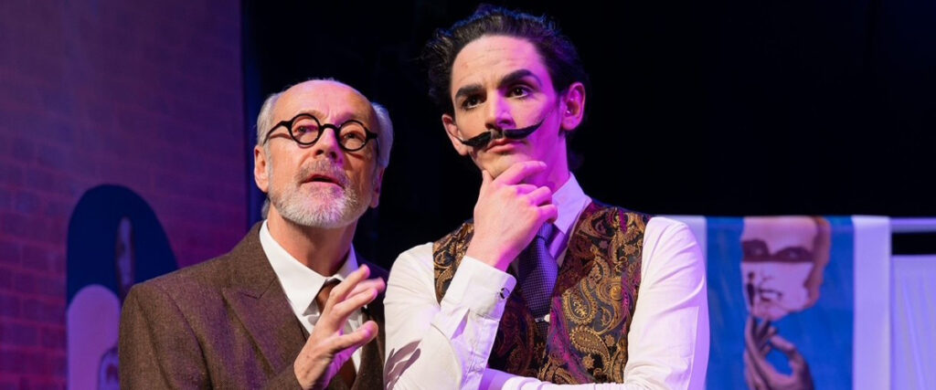 Two male characters in period costumes stand on stage. The older man with glasses looks intently, gesturing as if explaining. The younger man with a curled mustache ponders thoughtfully.