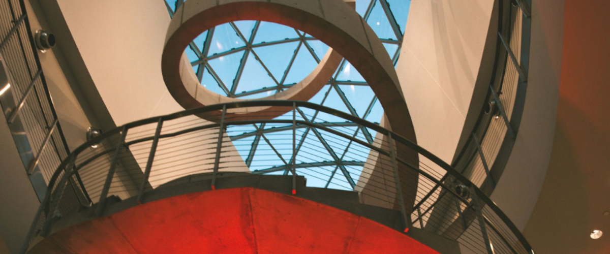 Upward view of The Dalí Museum staircase with geometric glass ceiling showing the blue sky, framed by curved railings and warm red lighting below.